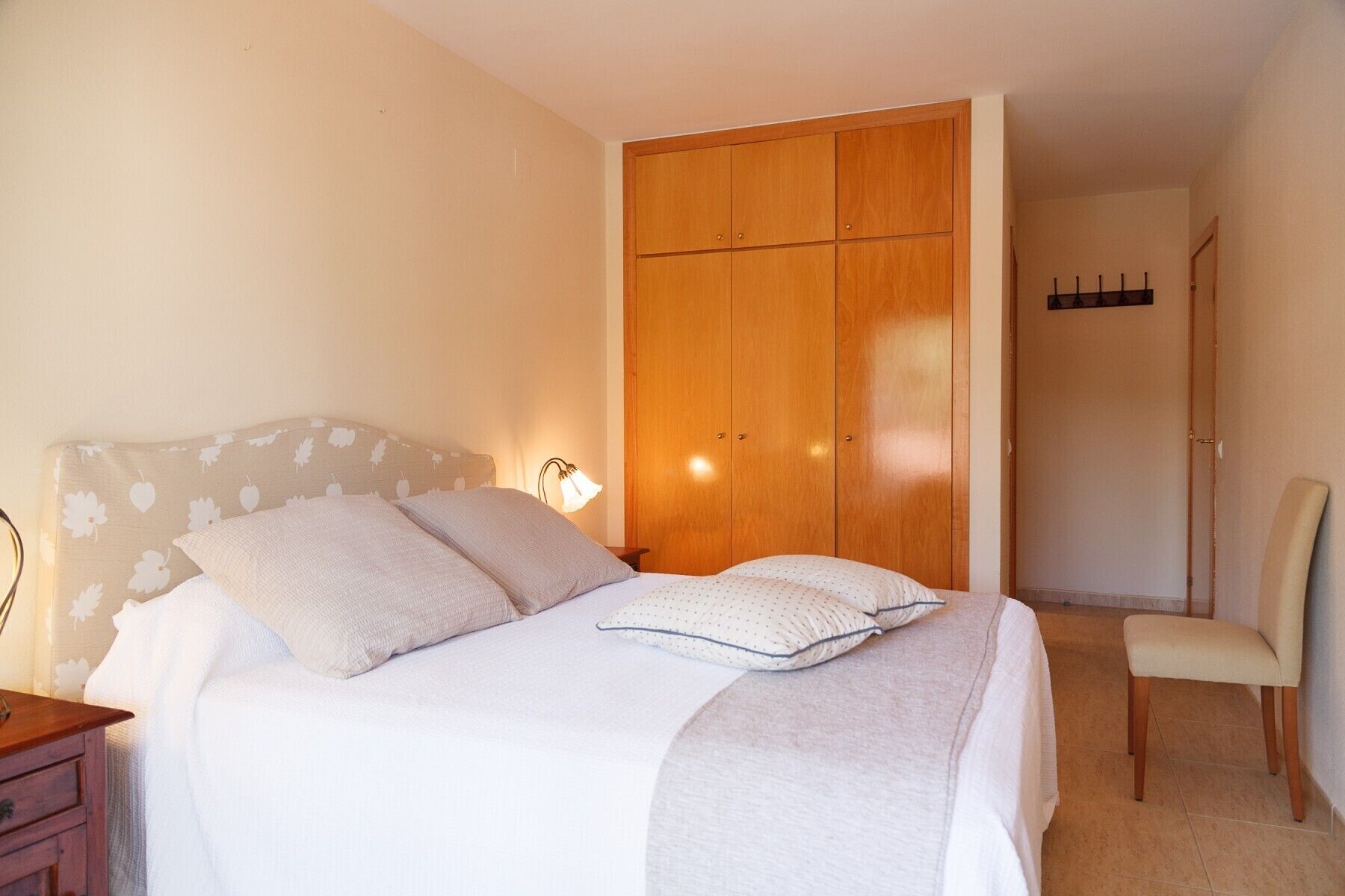 Un dormitorio con cama, mesita de noche, lámpara, armario y silla.