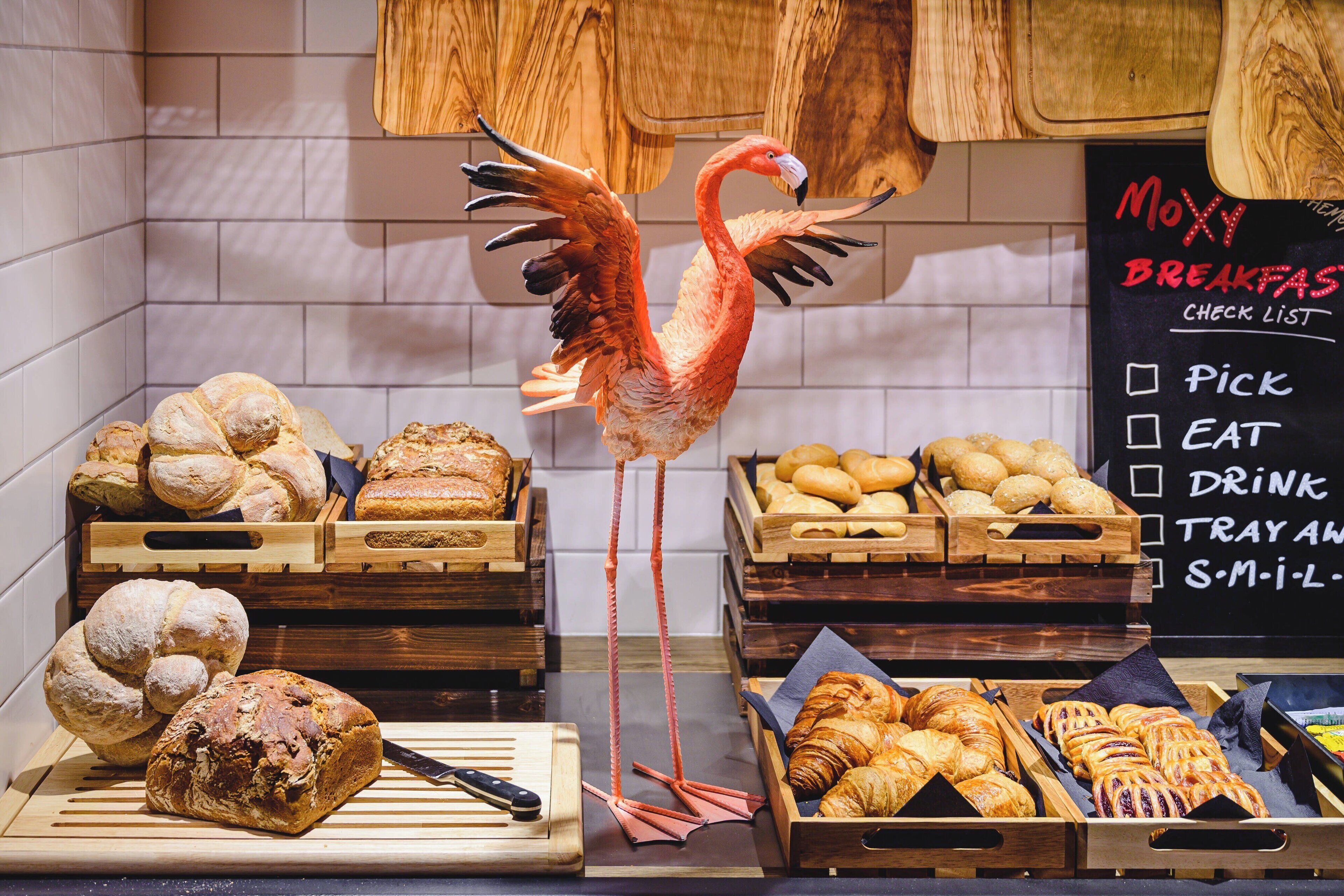 Un bufé de desayuno con panes y pastelería variados, una decoración de flamenco y un menú en pizarra que enumera las opciones de desayuno.