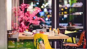 Een moderne eetruimte met een houten tafel, gele en zwarte stoelen, een gele koffer en een roze plant.
