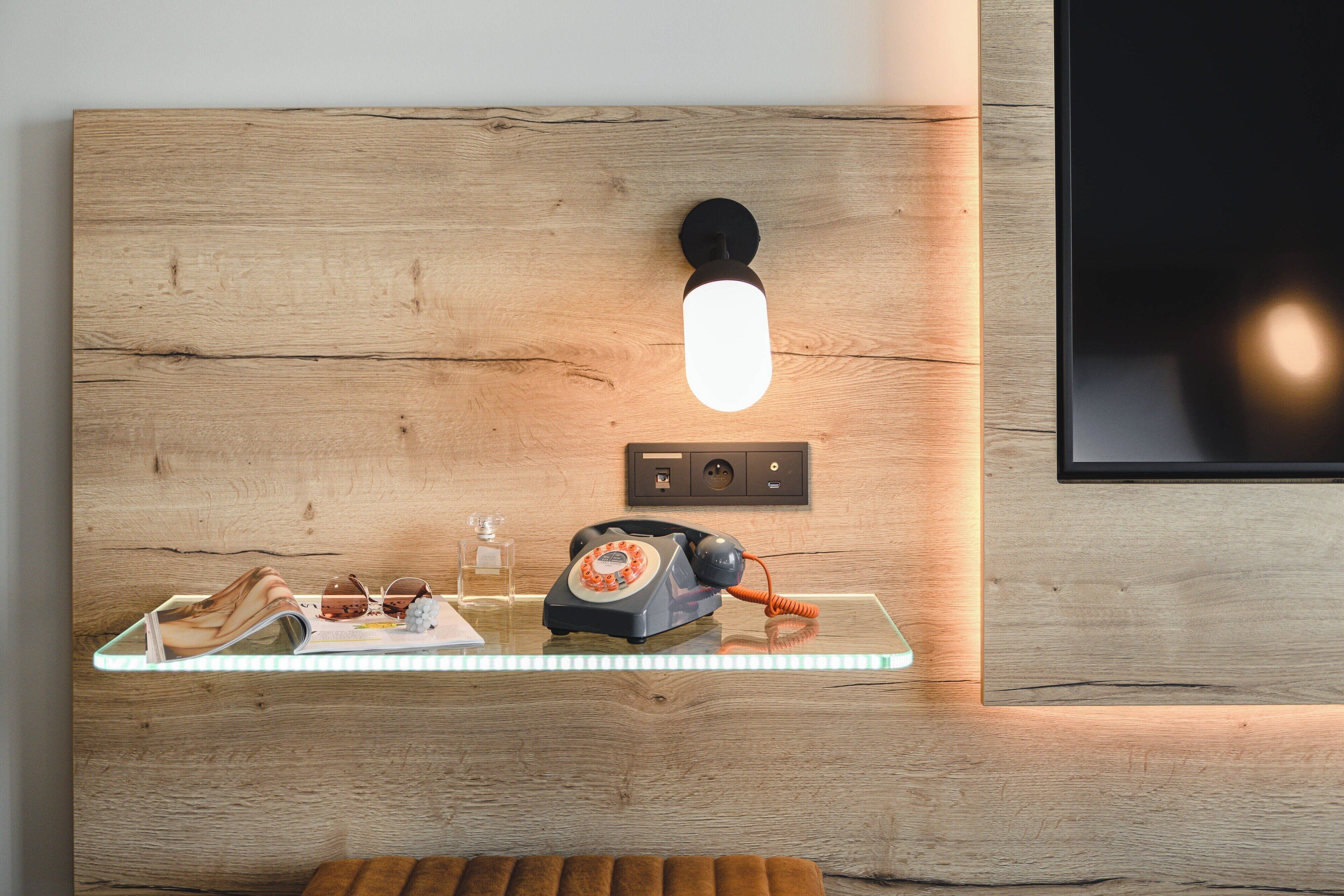 A wooden wall with a mounted telephone, a glass shelf with a magazine, and a small glass container.