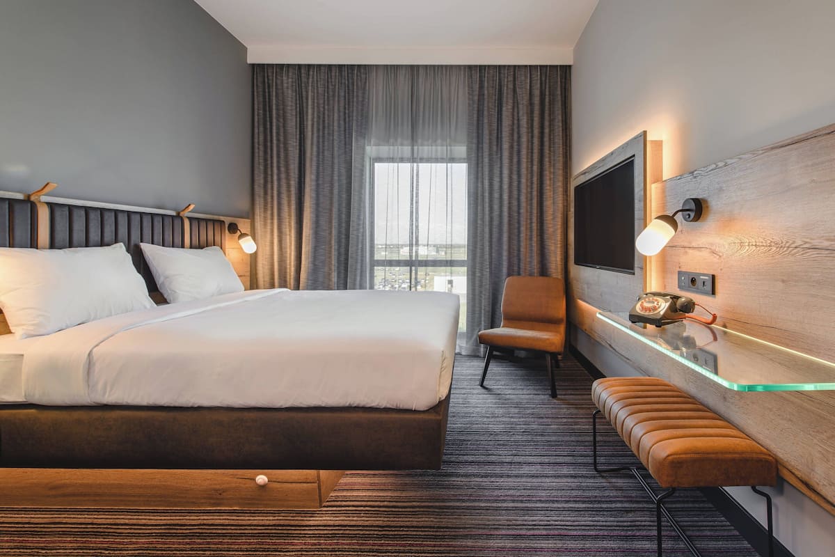 A hotel room with a large bed, a desk with a telephone, a chair, a bench, and a television on the wall.