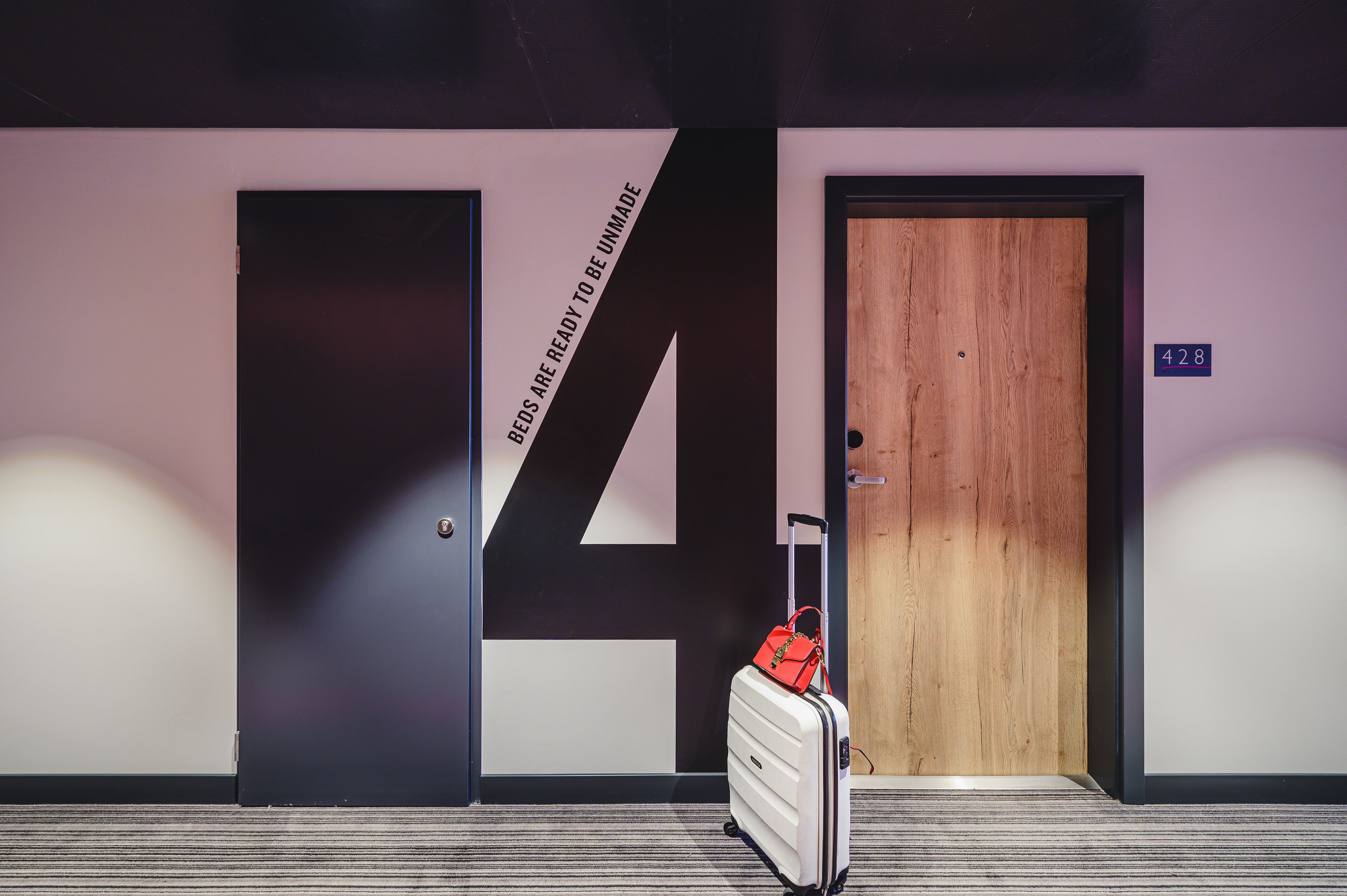 A hotel room with a large number '14' on the wall, a wooden door, and a suitcase with a red bag.