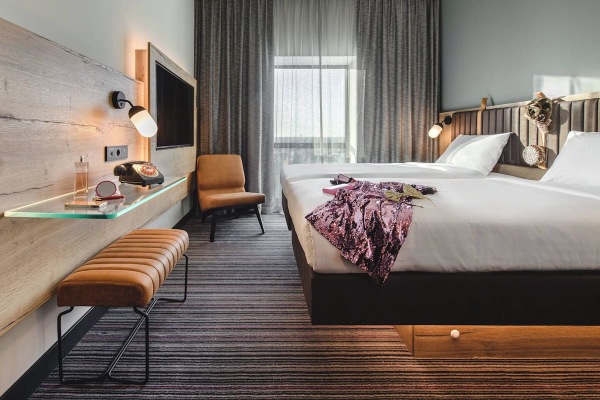 A modern hotel room with a large bed, a wooden headboard, a glass side table, a bench, a chair, and a television.