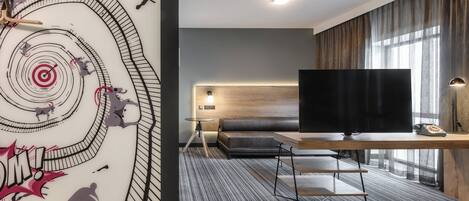 Una habitación de hotel moderna con una gran televisión de pantalla plana, un escritorio de madera, un sofá y una alfombra estampada.
