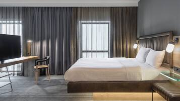 A modern hotel room with a large bed, a desk with a chair, a television, and a bench.