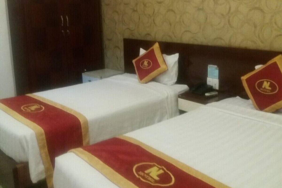 Ein Hotelzimmer mit zwei Betten, jeweils mit roter und goldener Bettdecke und einem Kissen mit einem Logo. Es gibt einen Holzschrank und eine an der Wand befestigte Ablage mit einem dekorativen Gegenstand.