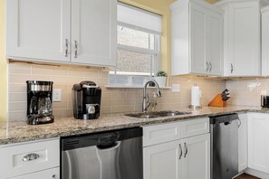 A modern kitchen with white cabinets, a granite countertop, a coffee maker, a dishwasher, and a sink.