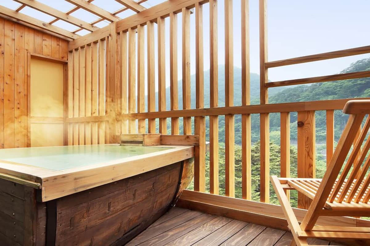 Um spa com sala de madeira, banheira de madeira, uma cadeira de madeira e vista para uma paisagem arborizada.