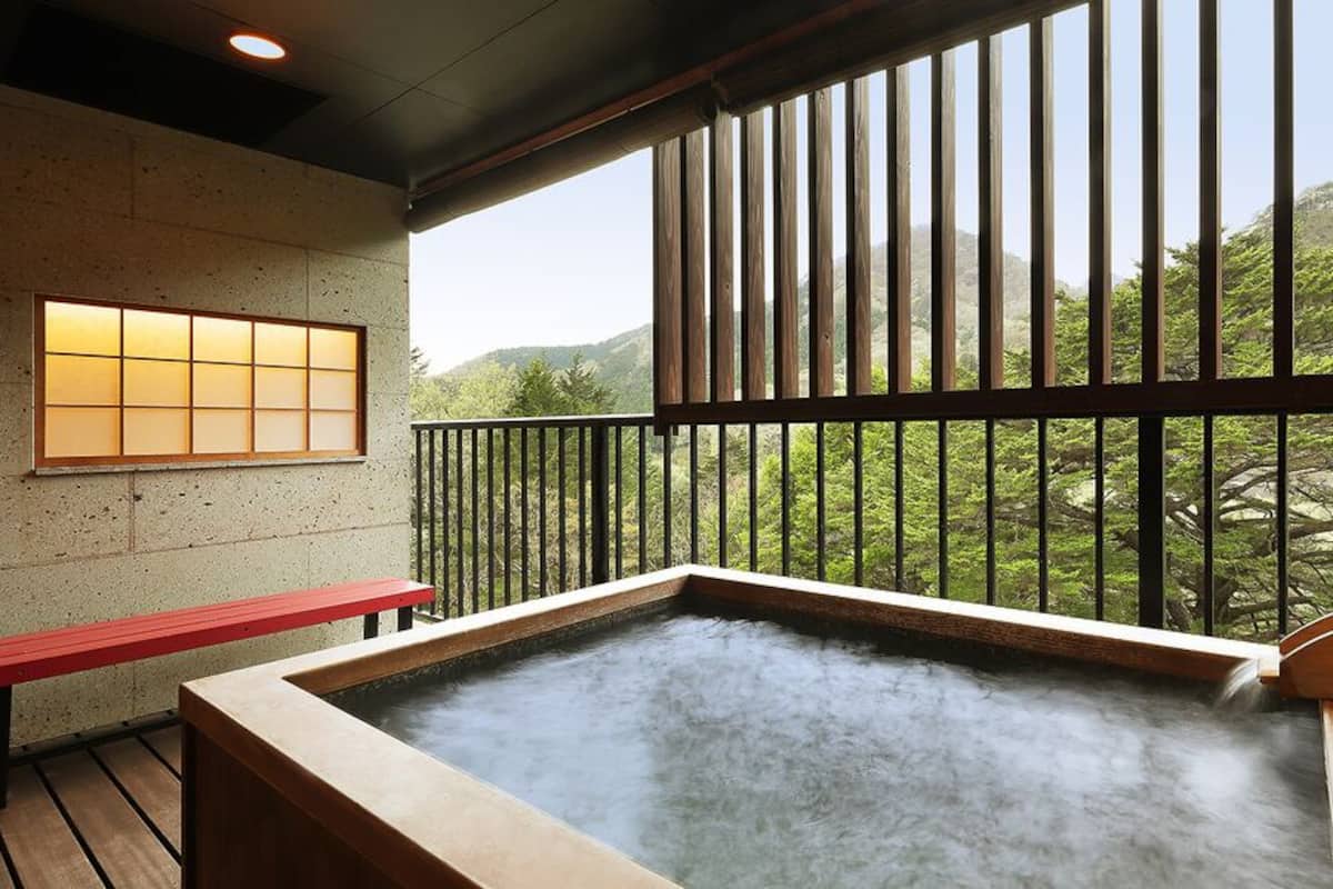 Uma área de águas termais com um banco de madeira, vista para o verde e uma janela tradicional japonesa.