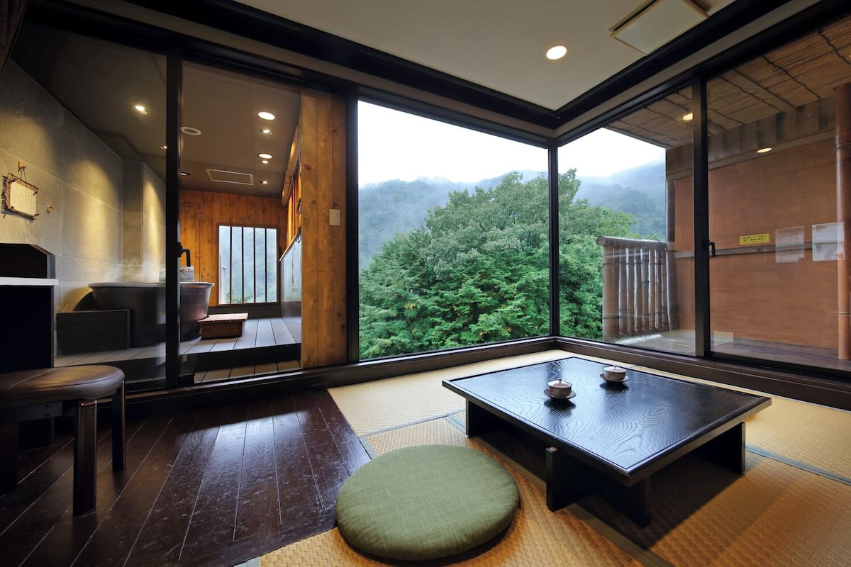 Um quarto japonês tradicional com tatames, uma mesa baixa com jogos de chá e vista para uma cadeia montanhosa arborizada.