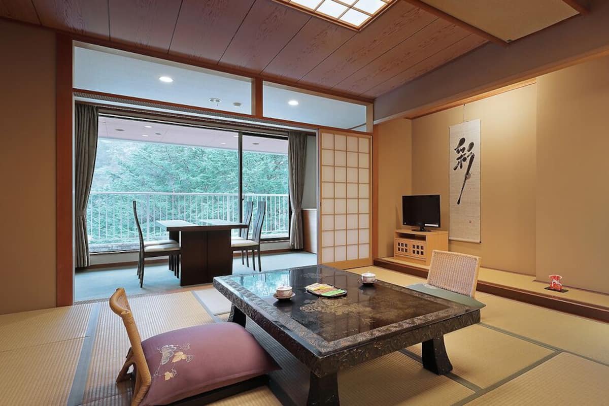 Um quarto japonês tradicional com piso de tatame, uma mesa baixa, um sofá, uma televisão e uma porta de correr que dá para uma varanda.