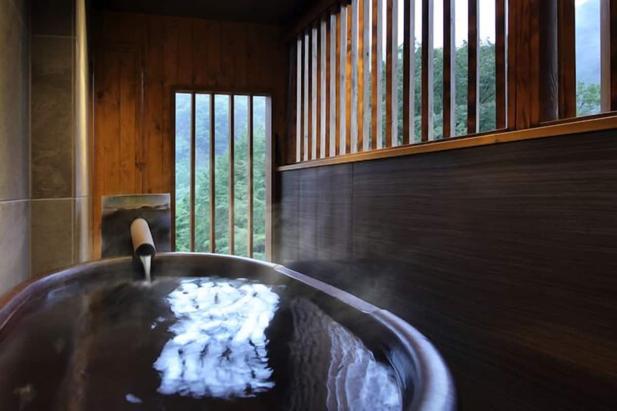 Uma banheira de hidromassagem com interior de madeira, uma janela com persianas verticais e vista para árvores.