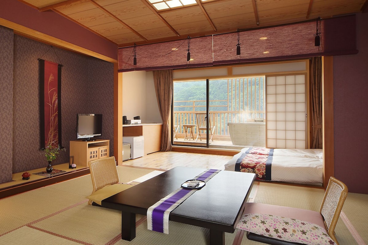 Quarto japonês tradicional com tatames, uma mesa baixa, televisão e uma porta deslizante para uma varanda externa.