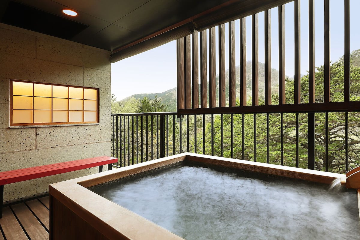 Uma área de águas termais com um banco de madeira, vista para vegetação e uma janela tradicional japonesa.