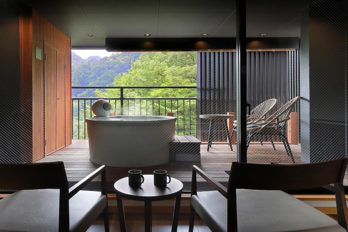 Quarto com banheira de hidromassagem, duas cadeiras, uma mesa e vista para o verde.