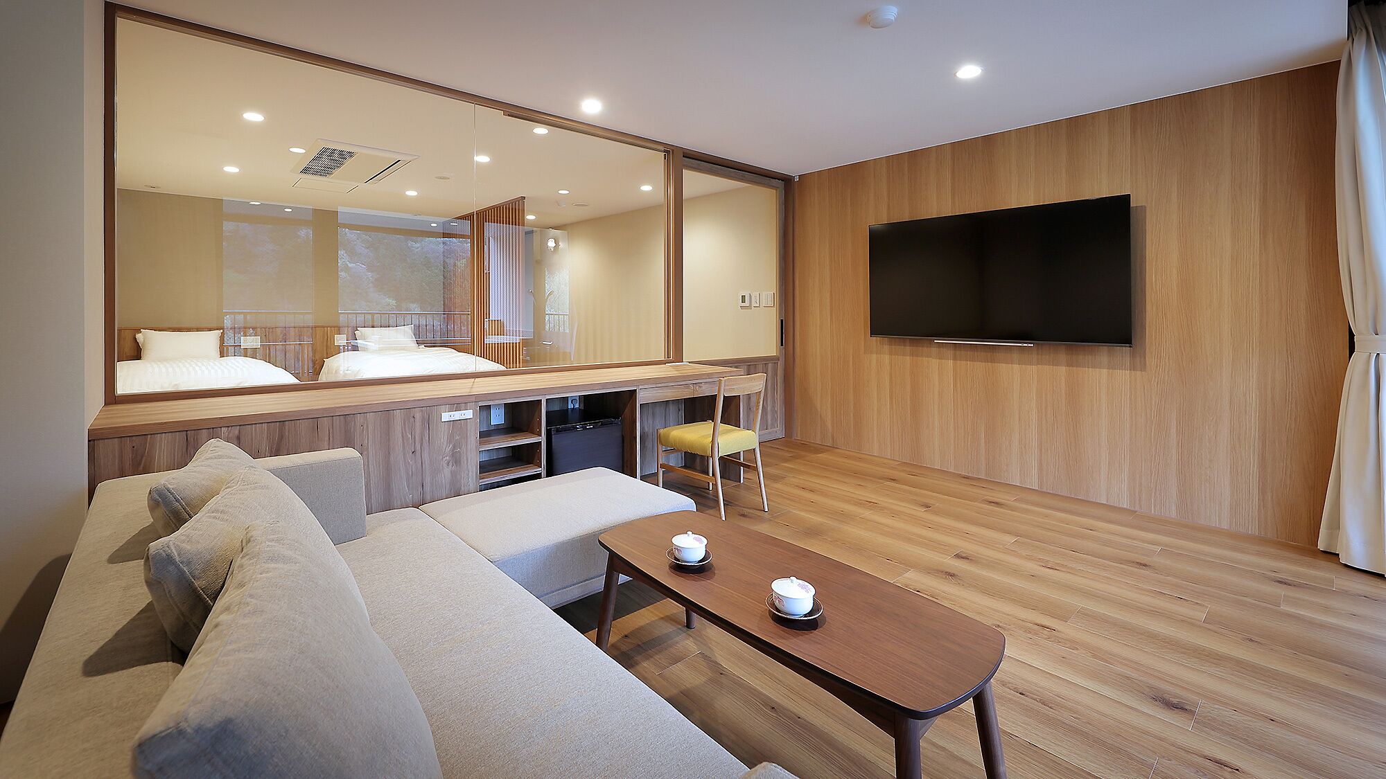 A modern hotel room with a sofa, a small table with cups, a TV, and a large mirror.