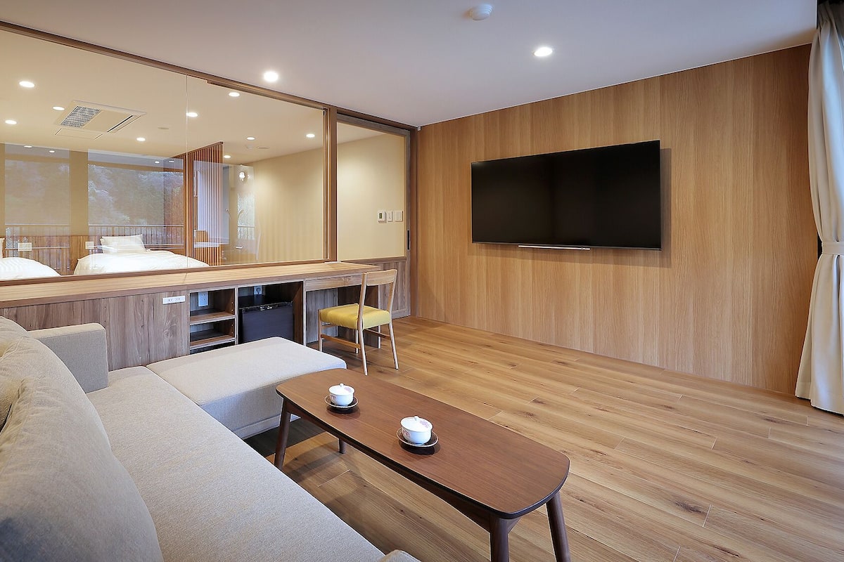 Quarto de hotel moderno com sofá, uma mesinha com chávenas, televisão e um espelho grande.