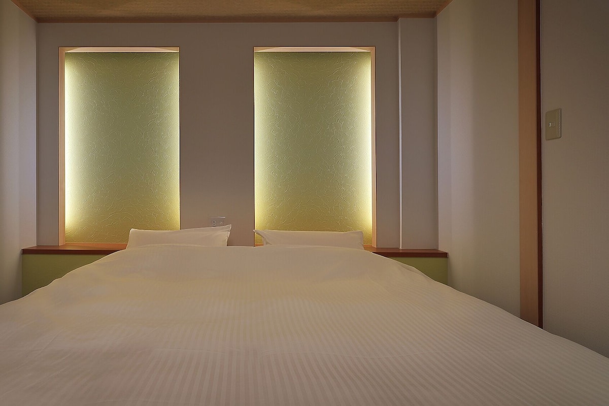 Quarto de hotel com uma cama grande, duas janelas com vidro texturizado e uma cabeceira de madeira.
