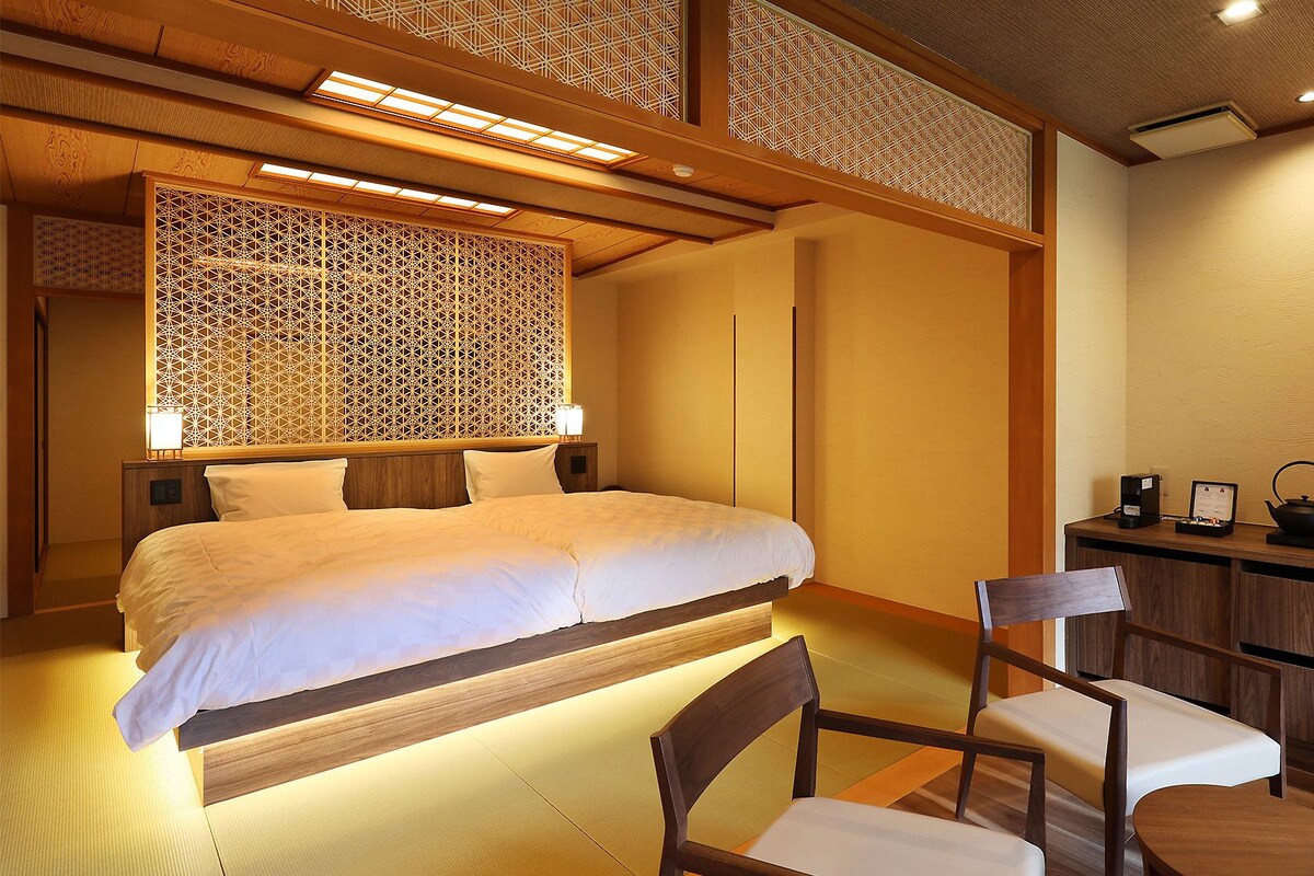 A hotel room with a bed, bedside tables, a chair, a desk with a kettle, and a small cabinet.