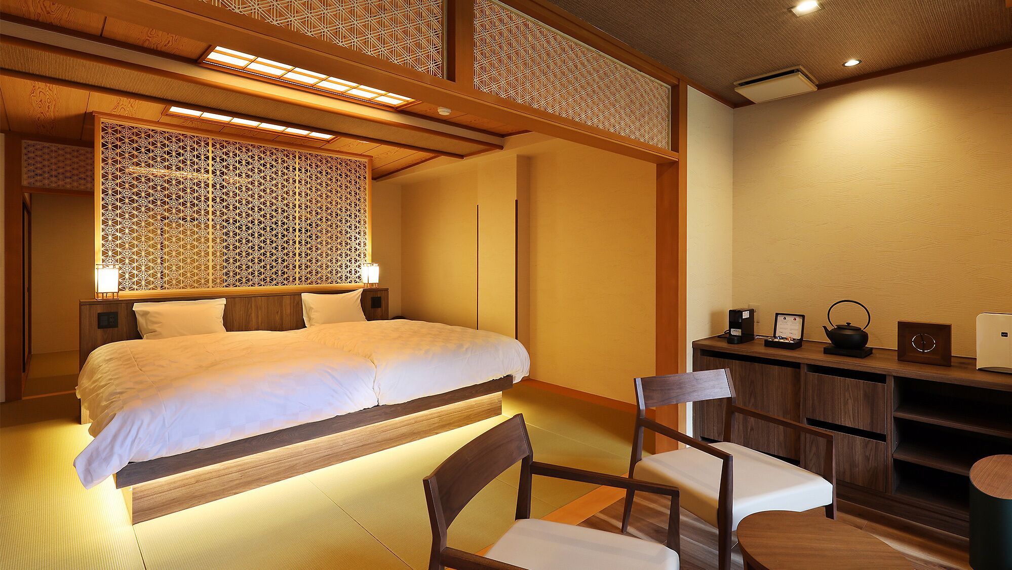 A hotel room with a bed, bedside tables, a chair, a desk with a kettle, and a small cabinet.