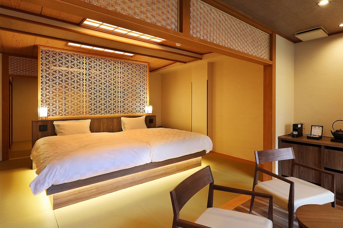 Quarto de hotel com cama, mesas de cabeceira, uma cadeira, escrivaninha com chaleira e um pequeno armário.