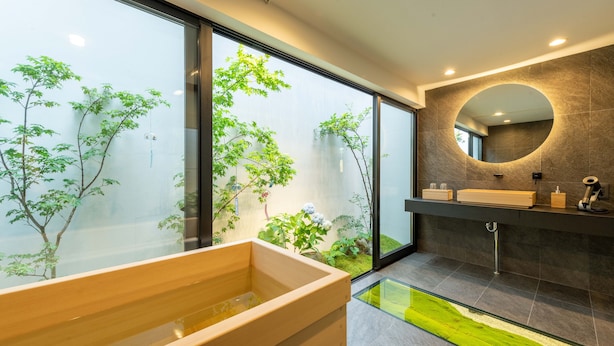 A modern bathroom with a wooden bathtub, a round mirror, and a view of greenery through large windows. - Tabitabi Stay Shinsen (Kyoto)