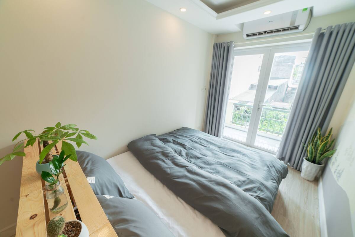 A bedroom with a bed, a wooden nightstand with plants, a window with curtains, and an air conditioning unit.