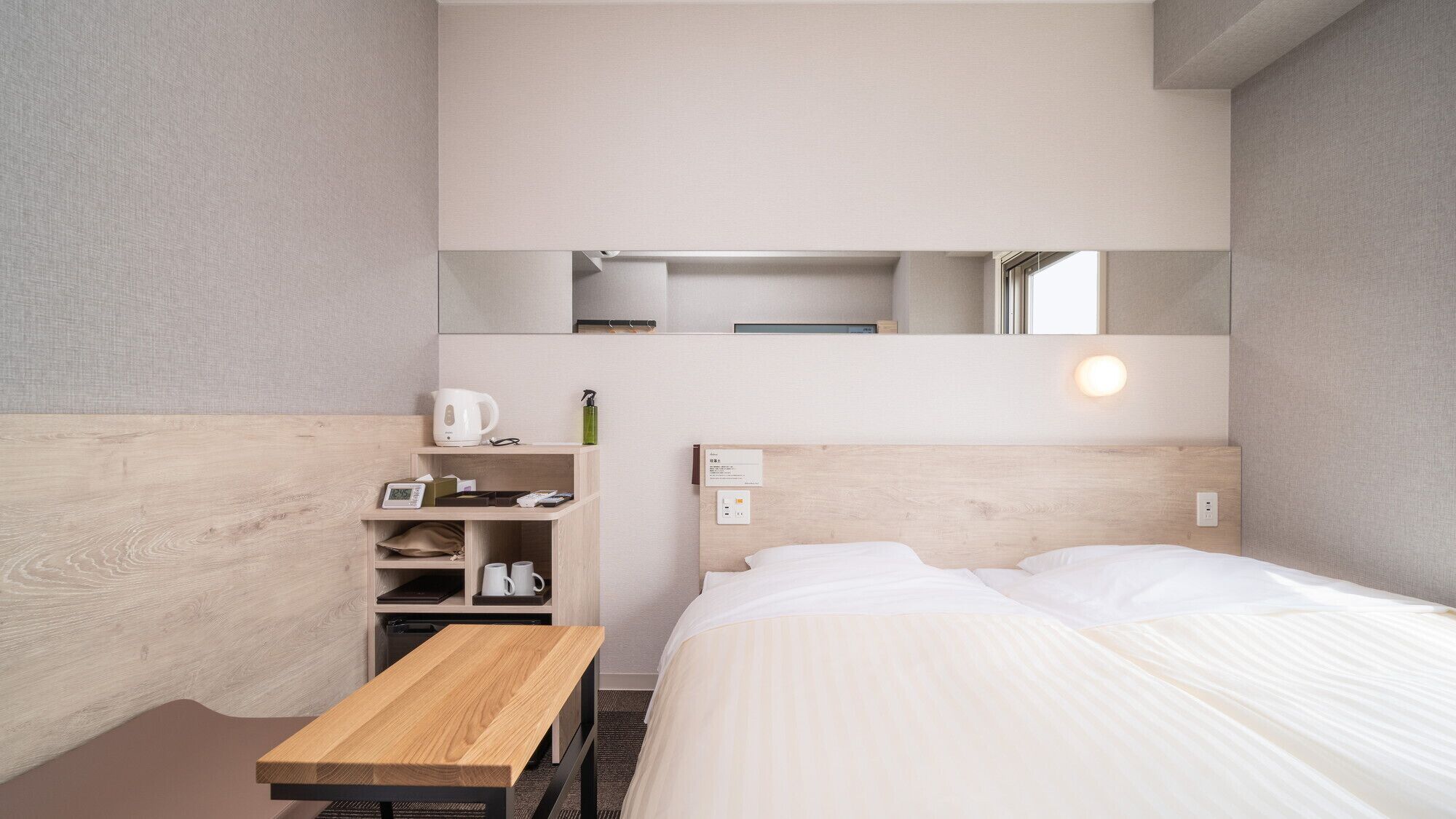Une chambre d’hôtel avec un lit, une table de chevet en bois, une petite table, une étagère avec des articles de toilette et un miroir au-dessus du lit.