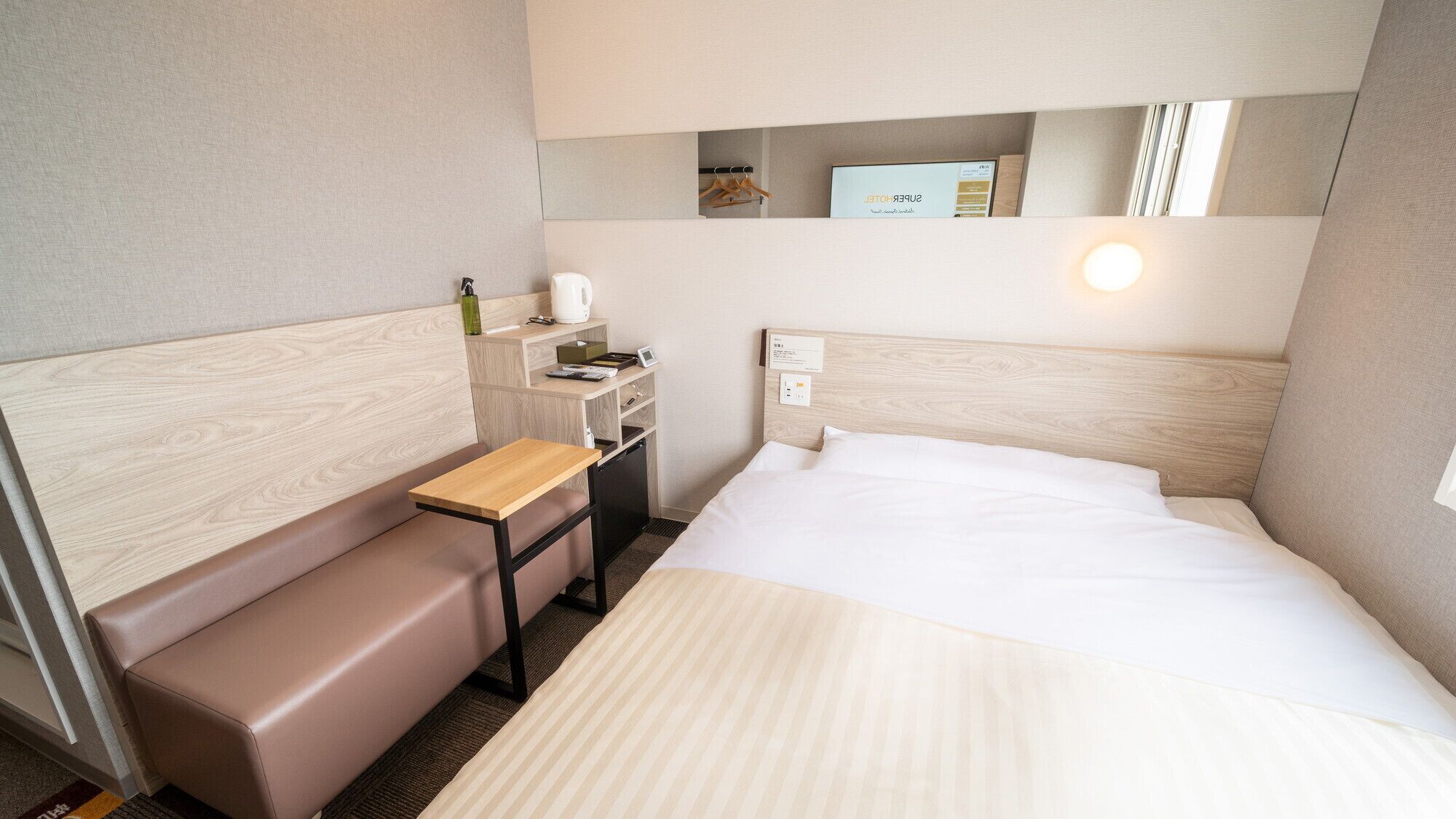 Une chambre d’hôtel comprenant un lit, un bureau, un banc, une petite étagère avec des articles de toilette et un miroir.