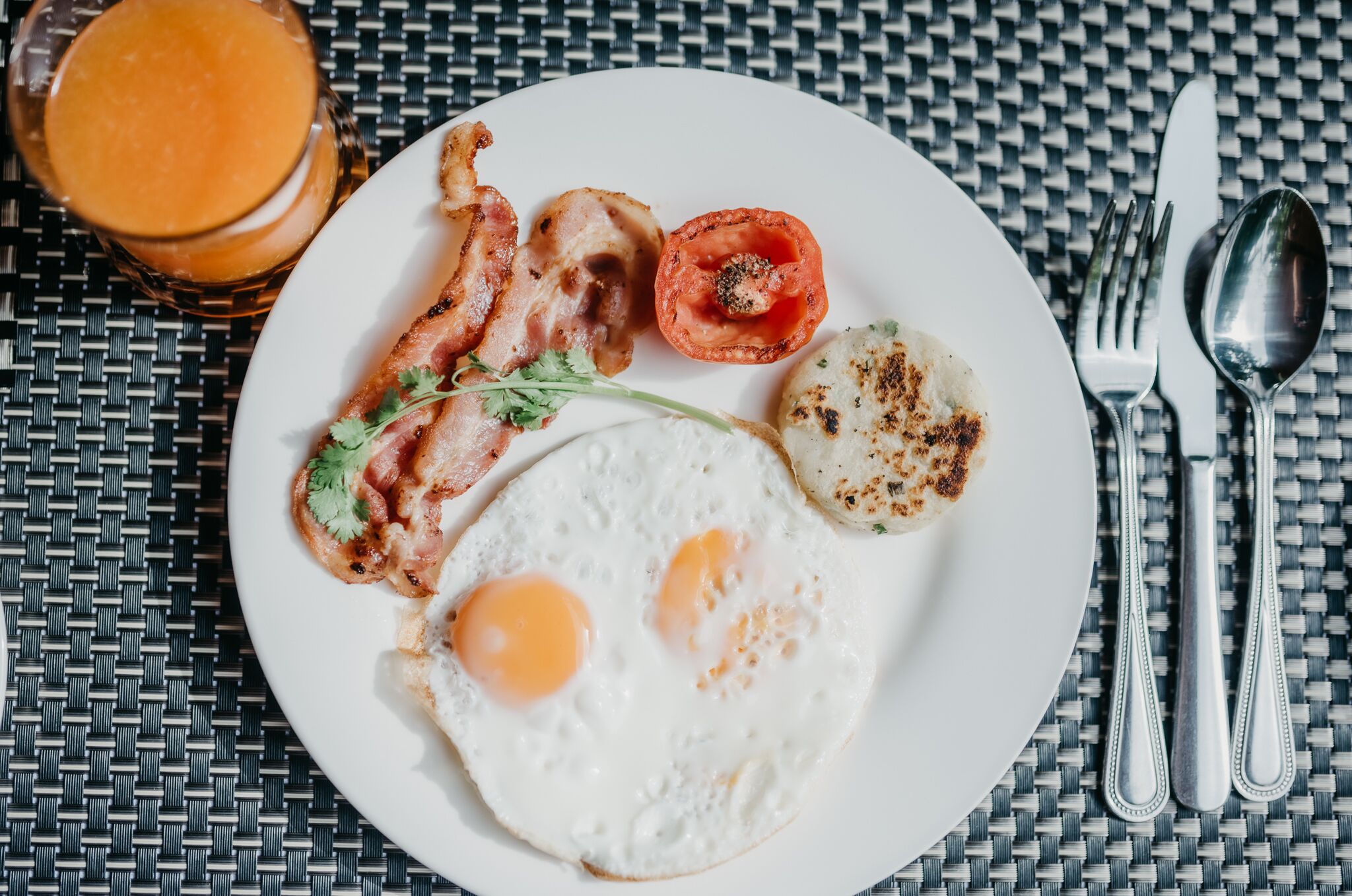 Une assiette de petit-déjeuner avec des œufs, du bacon, une tomate grillée et un verre de jus d’orange, le tout posé sur une nappe à motifs tissés.