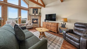A living room with a fireplace, a sofa, a leather chair, a television, and a large window. - Jackstraw Mountain 32 2 Bedroom Condo by RedAwning (Allenspark)