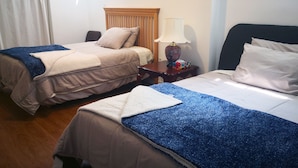 A hotel room with two beds, each with a blue and beige blanket, a wooden headboard, a nightstand with a lamp, and a small table with a vase and a box of tissues. - GA Stay Oakville Two Bedroom Guest House (Oakville)
