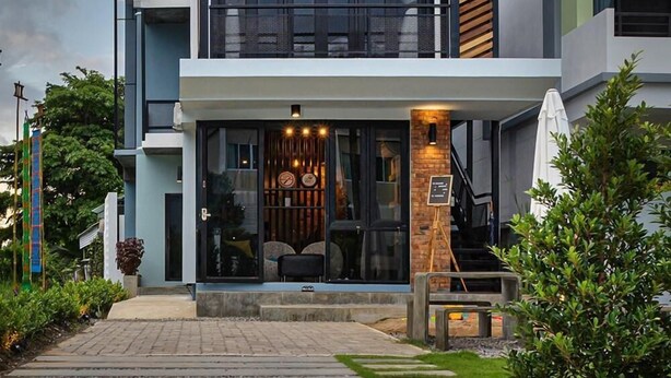 A modern two-story house with a balcony, large windows, and a well-lit entrance. - The Nichehome - A Lovely Waterside Living (San Sai)