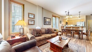 A living room with a sofa, coffee table, and a dining area with chairs and a table. There are windows with blinds, a lamp, and framed pictures on the wall.