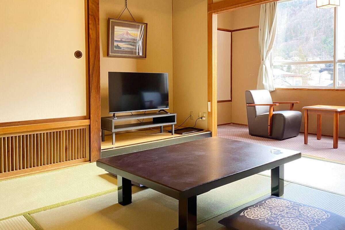A room with tatami flooring, a television, a table, chairs, and a window with a view.