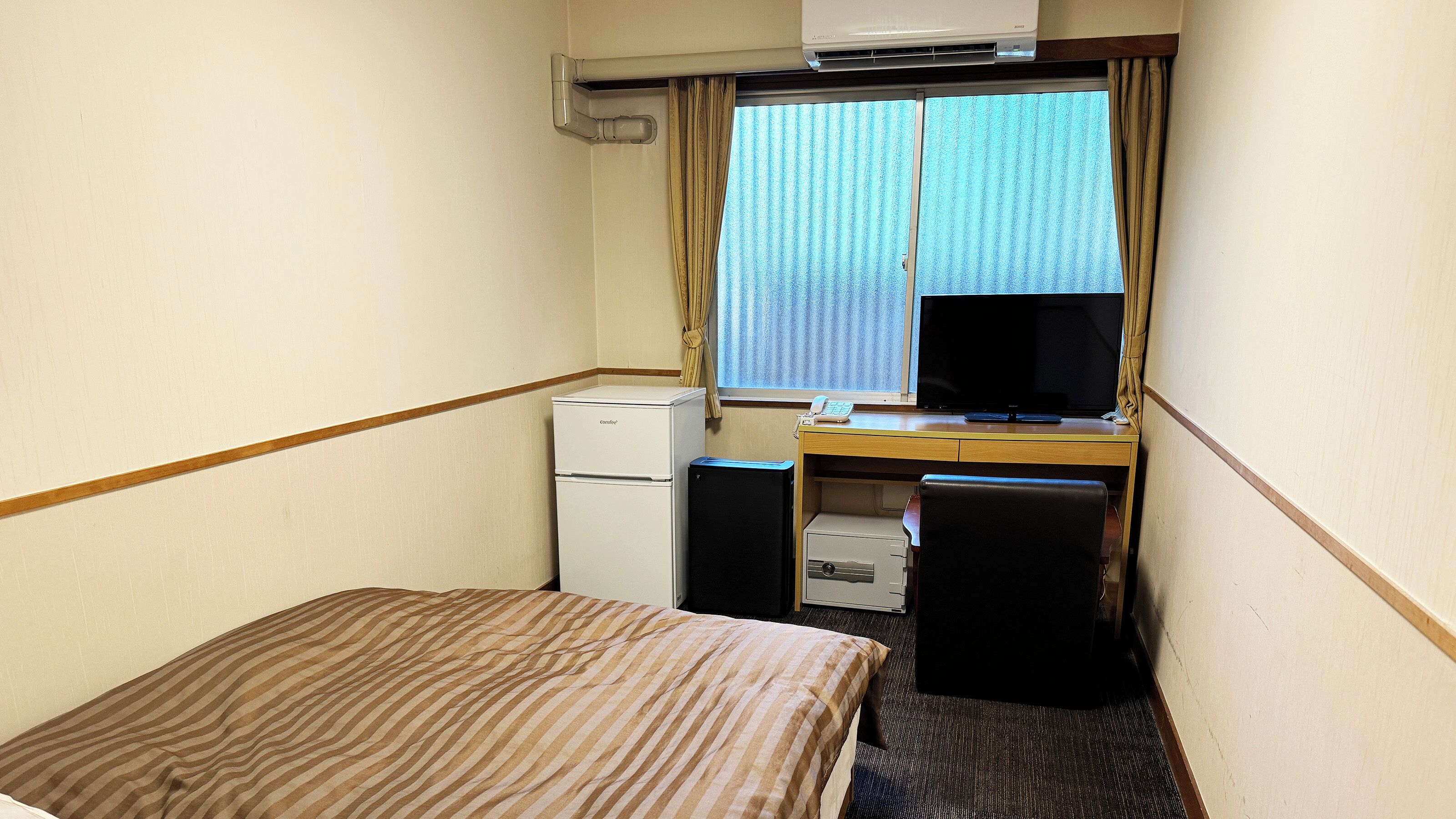 A compact hotel room with a bed, a desk with a computer, a small refrigerator, and a window with curtains.