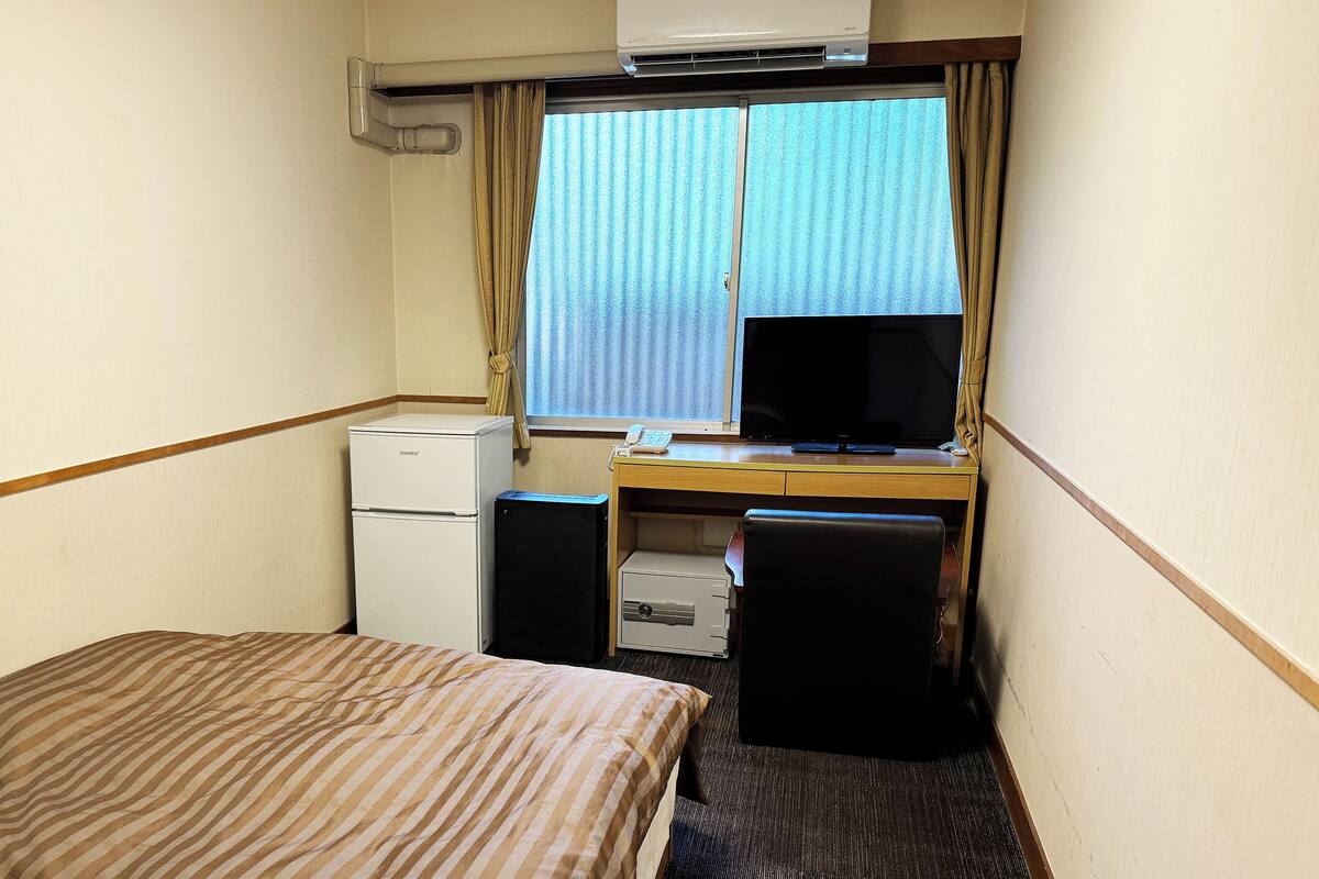 A compact hotel room with a bed, a desk with a computer, a small refrigerator, and a window with curtains.