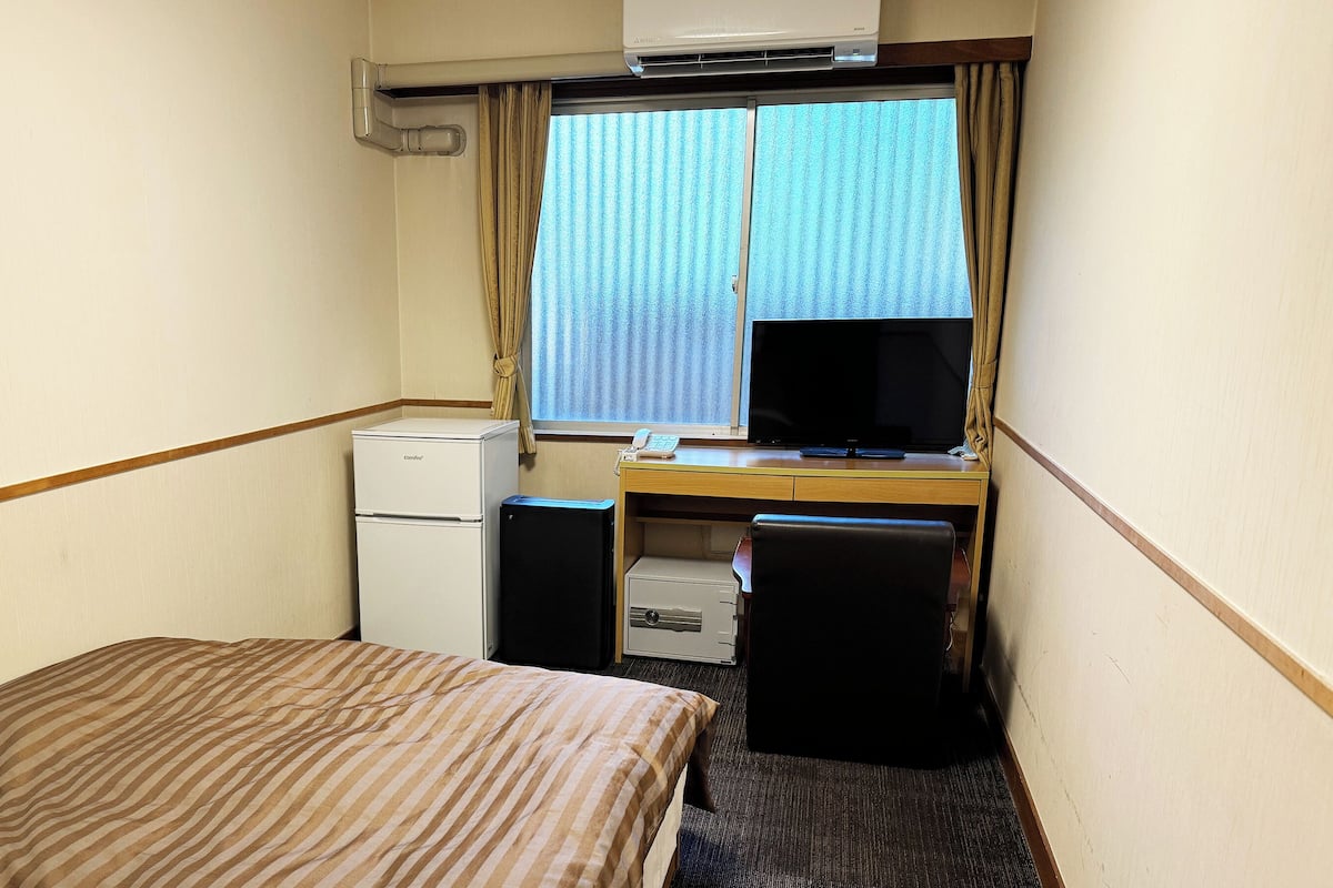 Ein kompaktes Hotelzimmer mit Bett, Schreibtisch mit Computer, kleinem Kühlschrank und Fenster mit Vorhängen.