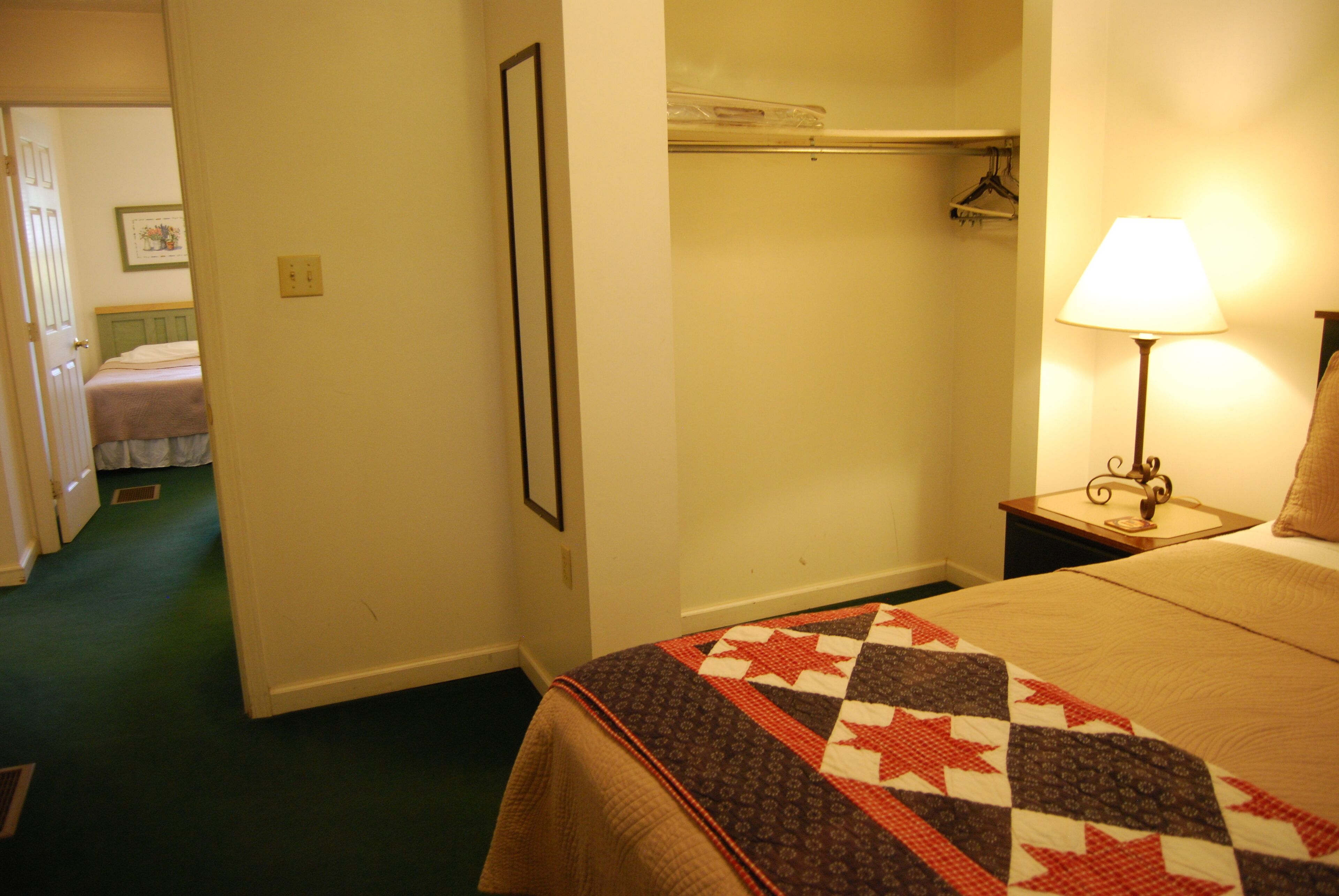 A hotel room with a bed, a nightstand with a lamp, a closet with a coat rack, and a door leading to another room.