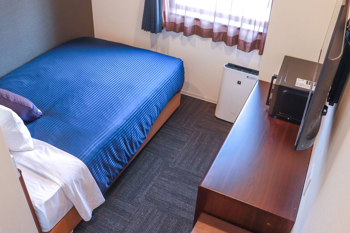 Ein kleines, ordentlich eingerichtetes Hotelzimmer mit einem Einzelbett, einem kleinen Schreibtisch, einem Fernseher und einer kompakten Mülleimer.