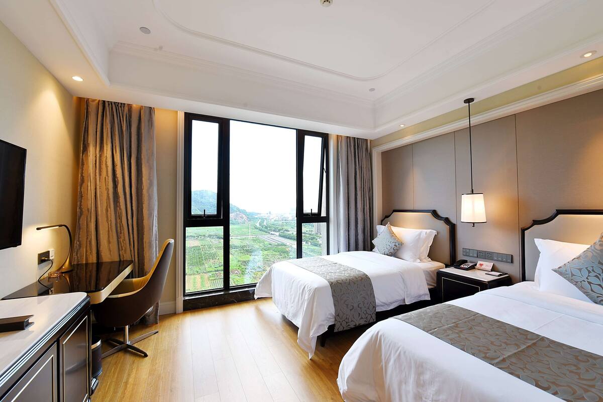 Ein Hotelzimmer mit zwei Betten, einem Schreibtisch, einem Stuhl und einem großen Fenster mit Blick ins Freie.