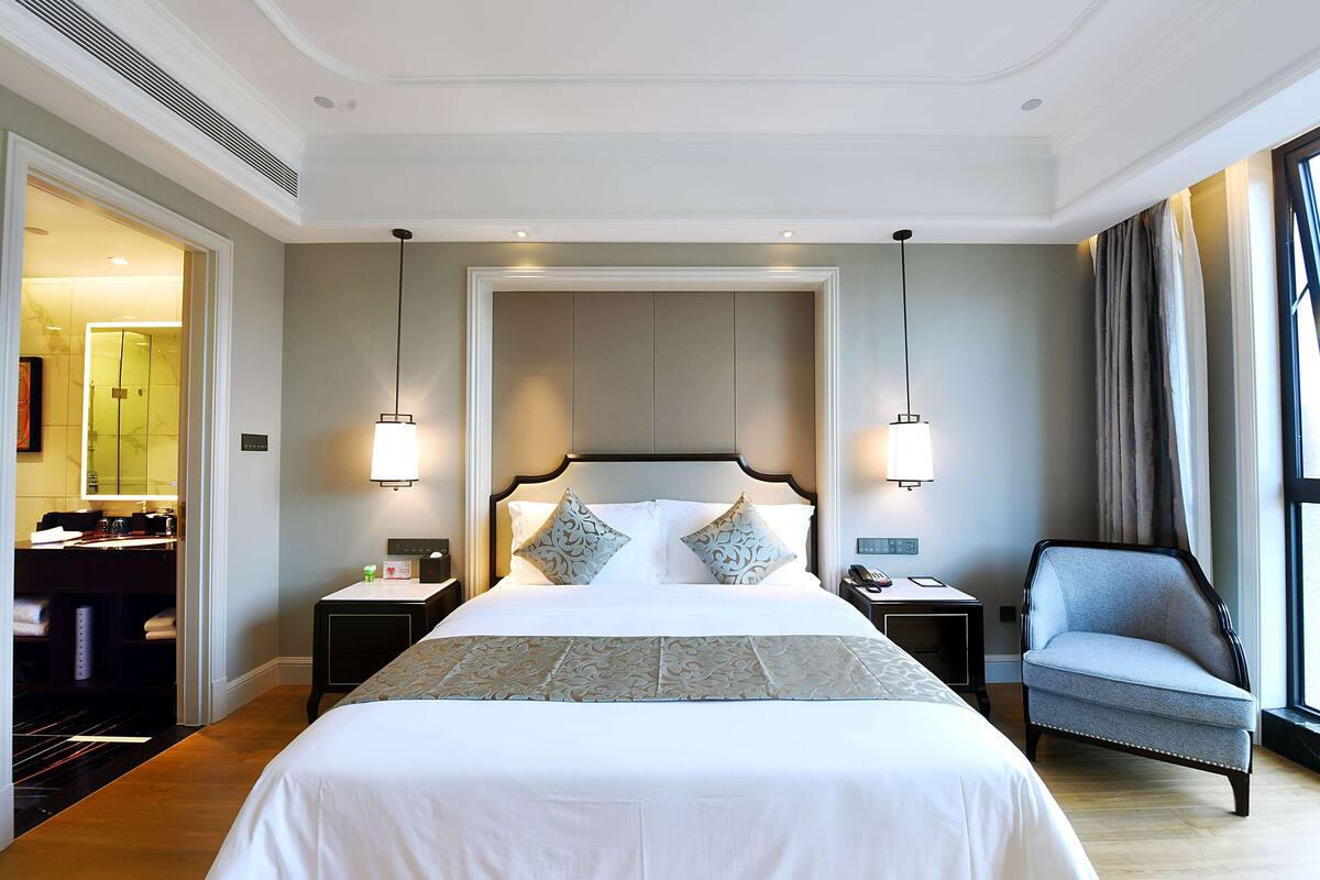 Ein Hotelzimmer mit einem großen Bett, zwei Nachttischen, einem Sessel, einem Schreibtisch mit Lampe und einem Spiegel.