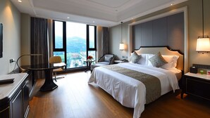 Une chambre d’hôtel avec un grand lit, un bureau, une chaise, une petite table ornée de fleurs et une vue sur la ville grâce à de grandes fenêtres.