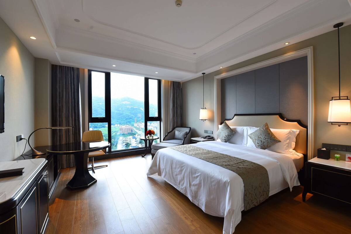 Ein Hotelzimmer mit einem großen Bett, einem Schreibtisch, einem Sessel, einem kleinen Tisch mit Blumen und Blick auf die Stadt durch große Fenster.