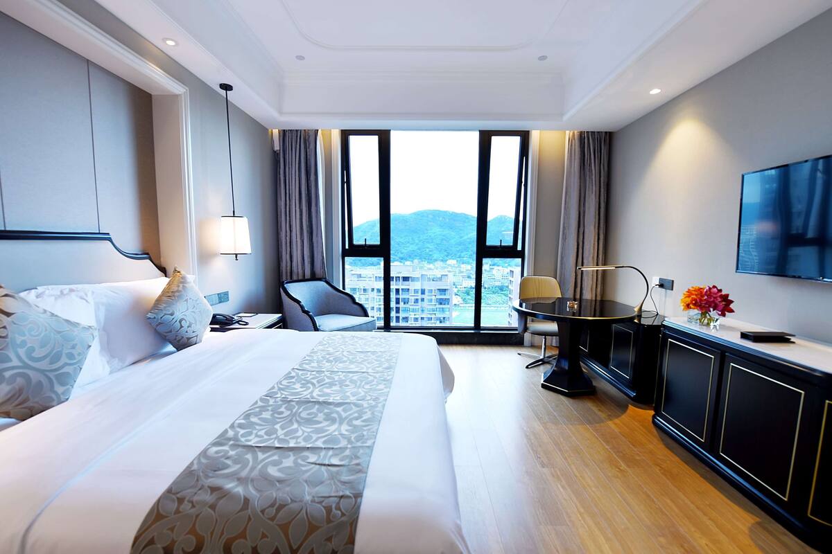 Ein Hotelzimmer mit einem großen Bett, einem Schreibtisch, einem Sessel, einem Fernseher und Blick auf eine Stadtlandschaft durch große Fenster.
