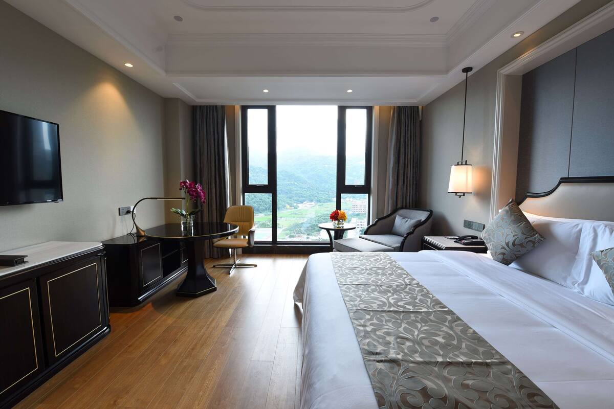 Ein Hotelzimmer mit einem großen Bett, einem Fernseher, einem Schreibtisch mit Stuhl, einem kleinen Tisch mit Blumen und Blick ins Freie durch große Fenster.