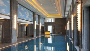 Une vaste piscine intérieure carrelée, entourée de murs dotés de grandes fenêtres et surmontée d’un plafond avec des luminaires encastrés.