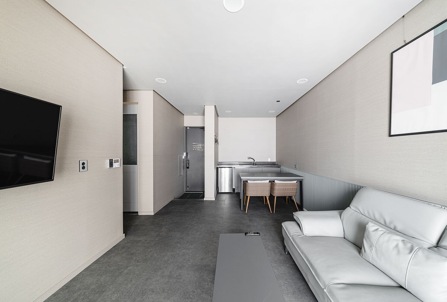 A modern, minimalist living room with a flat-screen TV, a sofa, a dining area with a table and chairs, and a kitchen area with a refrigerator and sink.