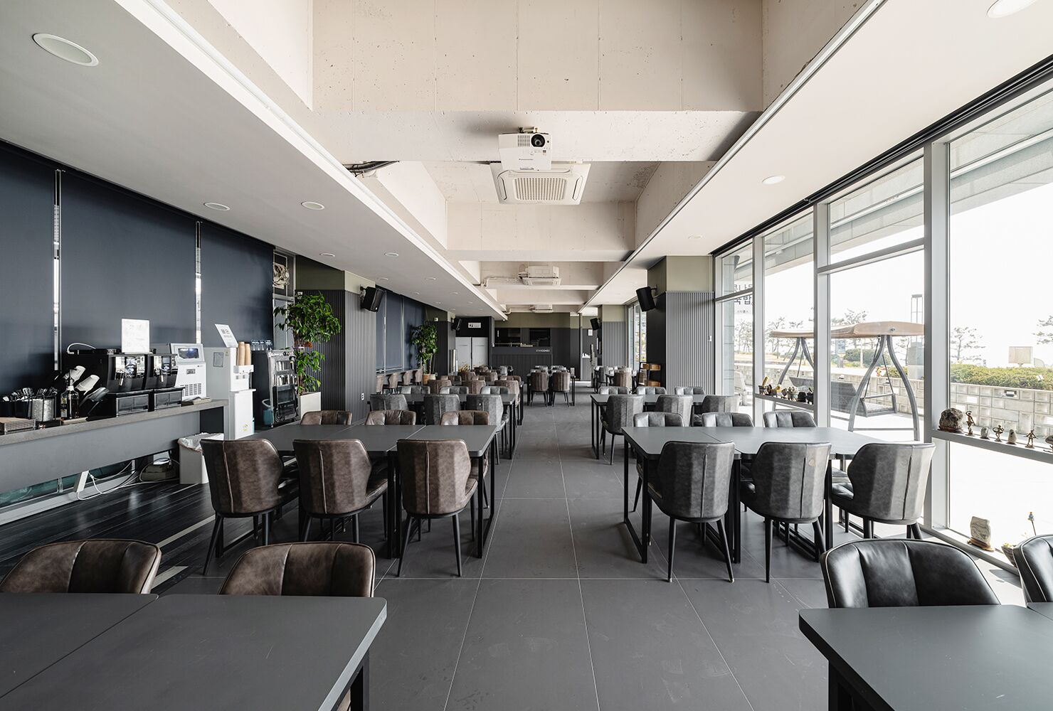 A modern restaurant with long tables and chairs, a kitchen area, and large windows offering a view.