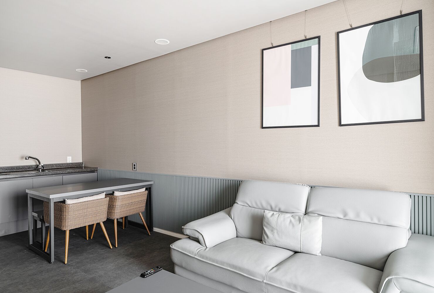 A modern living room with a grey sofa, a small dining table with wicker chairs, and a kitchen area with a sink and countertop.